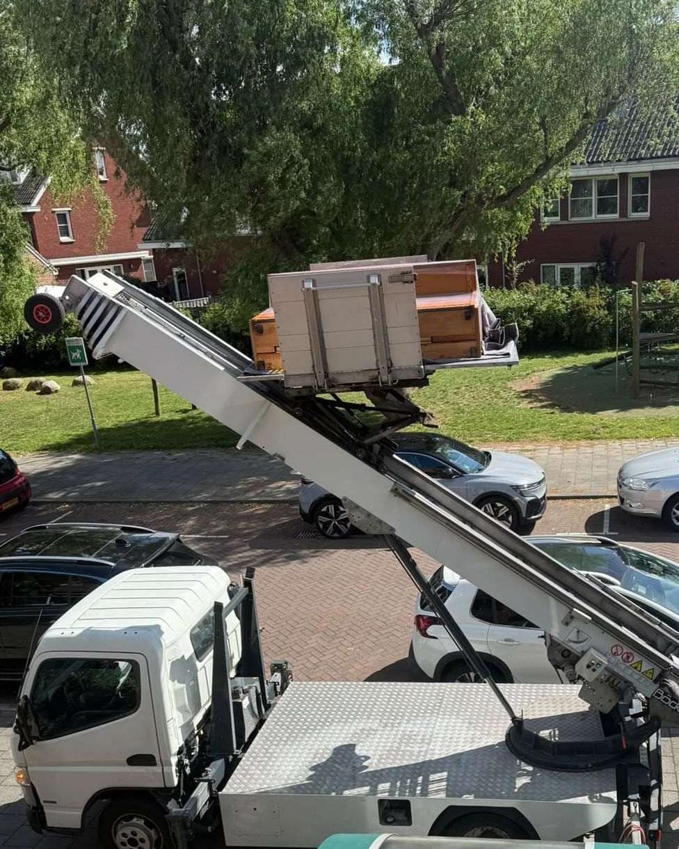 Piano op het platform van een verhuislift, onderweg naar een verdieping, wagen met laadklep ervoor