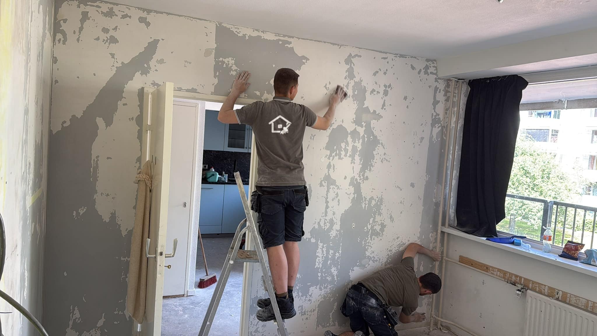 Twee medewerkers halen in een leeggeruimde woning de wandafwerking eraf, met een trap en gereedschap op de kale vloer